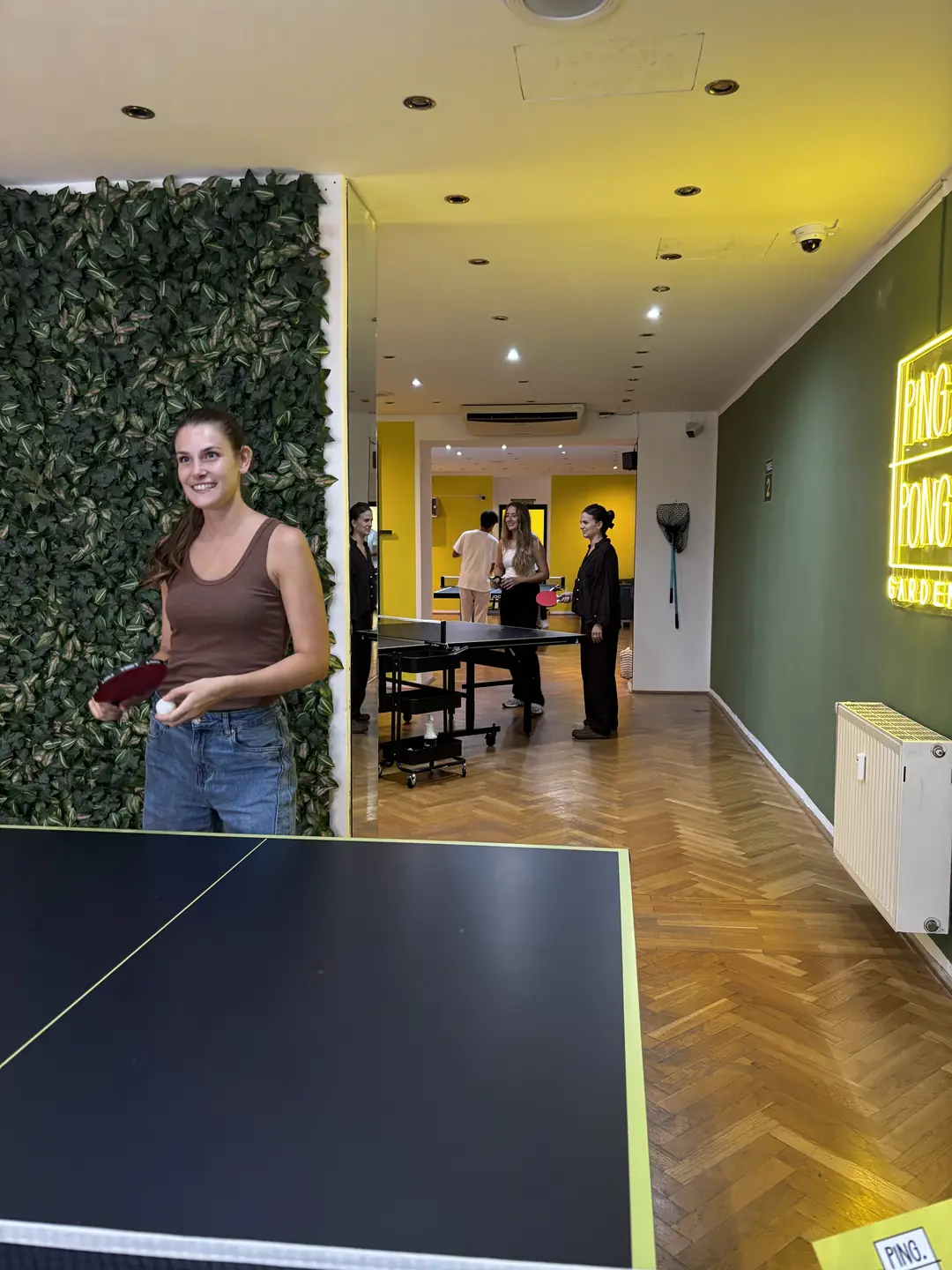 Spielerin mit Schläger vor der Pflanzenwand, dahinter der Gang zu den weiteren Tischen