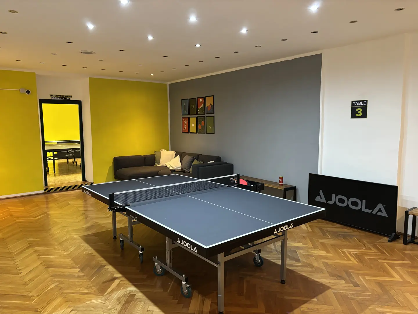 Heller Spielraum mit Tischtennisplatte an Tisch 3, Lounge-Sofa und Fischgrätparkett